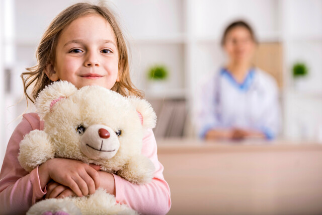 A photo. A child in a doctor's office, smiling and holding a teddy bear, a doctor in the background.
