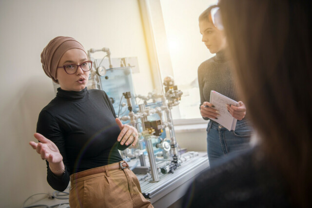 Photo of a person read as female standing in front of other people in a laboratory and explaining something.
