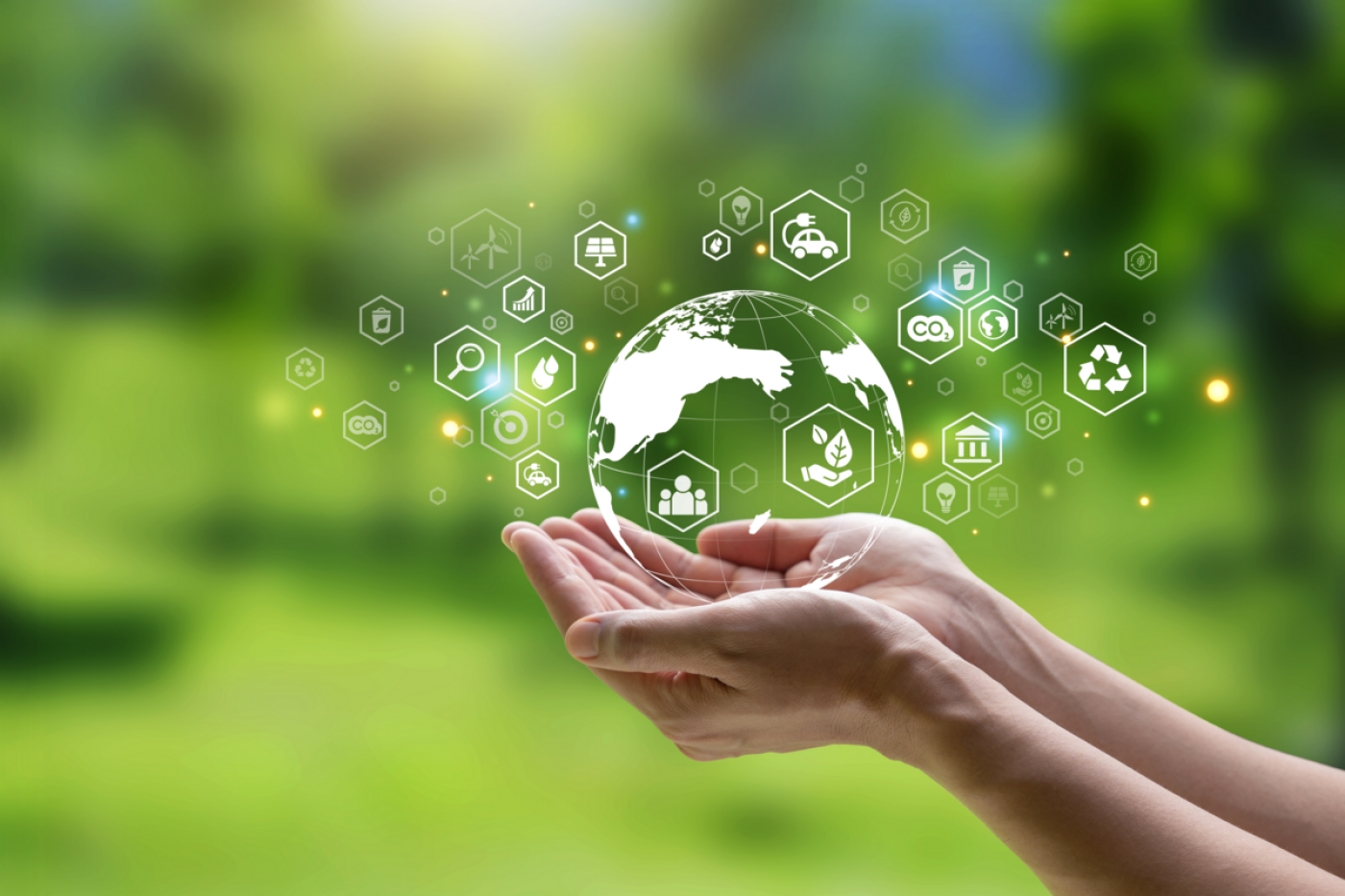 Two hands hold a transparent globe. Surrounding the globe are small hexagons with drops of water, recycling arrows, wind turbines and much more. A blurred background with greenery.