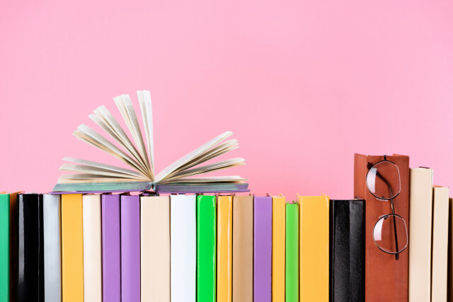 The photo shows a row of books against a pink background. One book lies open on top of the others. Glasses are hanging from another book. 