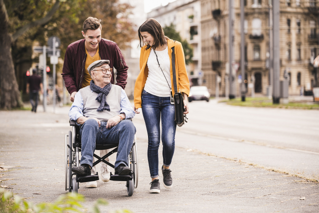 Das Foto zeigt einen Mann und eine Frau, die mit ihrem, im Rollstuhl sitzenden, Großvater spazieren gehen.