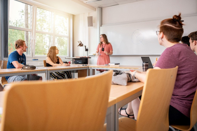 Das Foto zeigt ein paar Studierende in einem Seminarraum, während die Dozentin etwas erklärt.