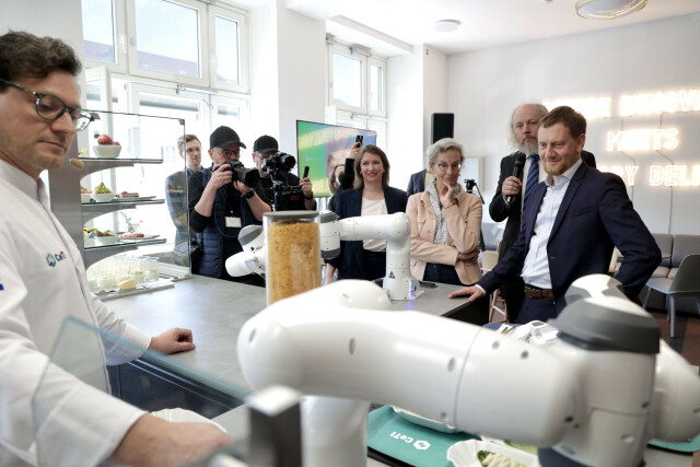 Eine Gruppe von Menschen beobachtet Roboterarme, die einem Koch in einer Bar helfen.