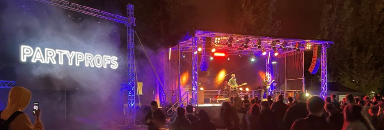 In lila-blau beleuchtete Bühne auf dem Campusfest. Spielende Band vor Publikum. Leuchtschriftzug 'Partyprofs' links.