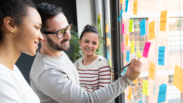Drei Personen stehen vor einem Fenster mit verschiedenfarbigen Post-Its. 
