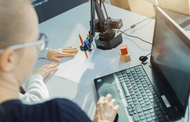 Over-the-Shoulder-Shot. Zu sehen sind zwei Personen, die am Laptop sitzen, sowie ein Roboterarm, der mit einem Bleistift zeichnet.