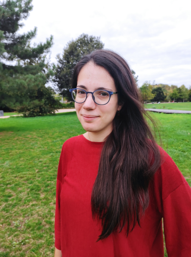 Clara Renedo, a woman with long dark hair and glasses, is standing outside on a grassy lawn. She is wearing a red pullover.