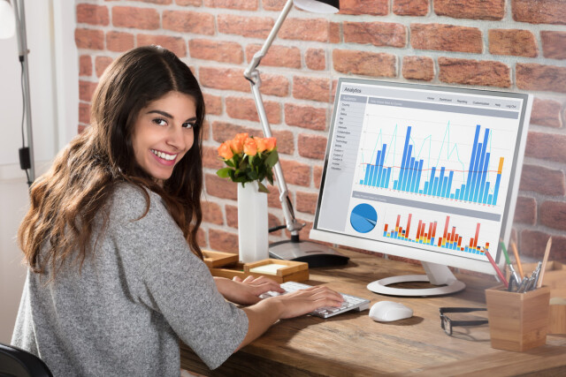The image shows a young woman. She is working on her computer and smiling at the camera. 