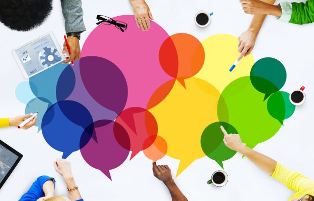 A white rectangular tabletop can be seen from above, on which different colored and differently sized round speech bubbles lie in the middle. The arms and hands of 7 different people can be seen around the tabletop.