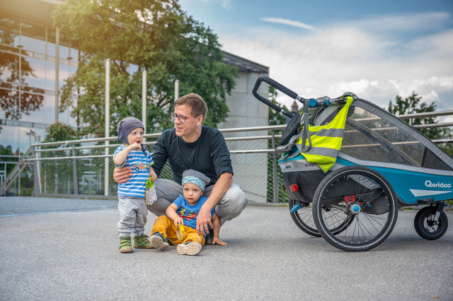 The photo shows a father with his two children. Next to them is a bicycle trailer for children.