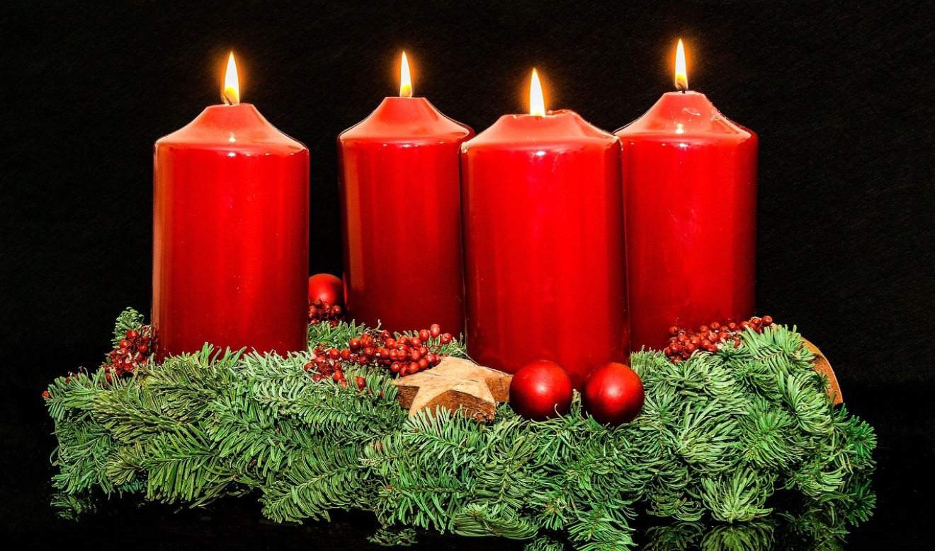 Ein Adventskranz mit vier großen, roten Kerzen, von denen alle brennen. Der Kranz ist aus Tannenzweigen gefertigt und mit roten Beeren, kleinen Kugeln und einem Stern dekoriert.