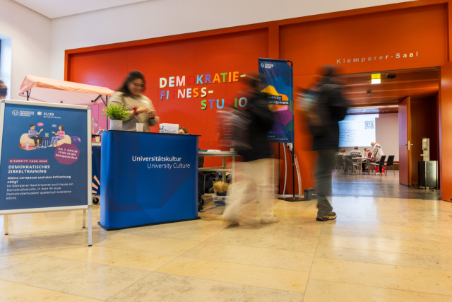  The photo shows a person in a room behind a counter. “University Culture” is written on the counter. Two people are walking past the counter in the photo.