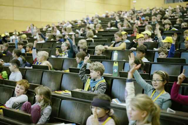 look into a lecture hall where a lot of children are sitting, some of them are raising their hands