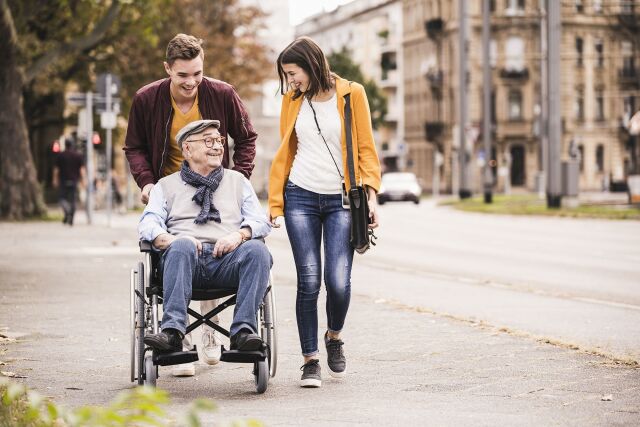 The picture shows an older gentleman in a wheelchair turning to a young woman next to him, while a young man pushes the wheelchair.