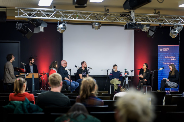 Six people are seated in a semicircle around a table, engaged in a lively discussion. The atmosphere is focused and interactive, with some participants gesturing while speaking and others listening attentively.