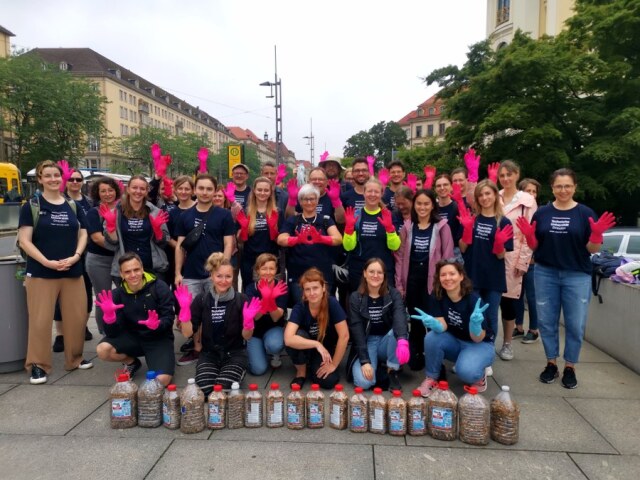 About 35 people in blue t-shirts stand in 4 rows one behind the other, holding up their hands in pink gloves. In front of them are 18 large plastic bottles filled with cigarette butts.