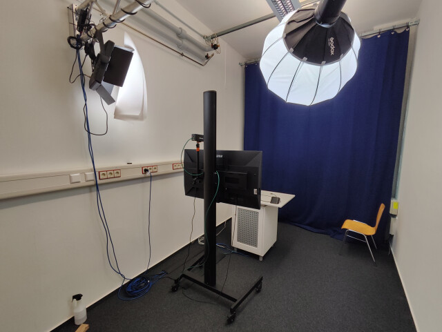 Kompakter Videoraum mit Kamera, Softbox-Beleuchtung, Monitor, Tisch und dunklem Vorhang im Hintergrund.