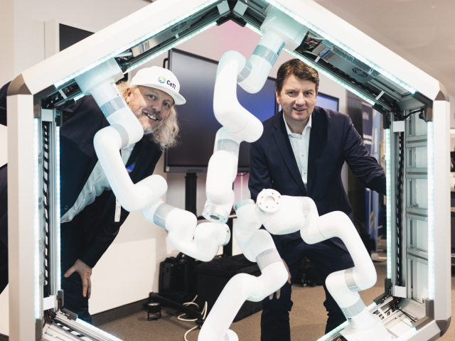 The picture shows two men stand behind a demo with robotic arms and look into the camera.
