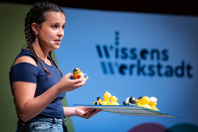 A young woman stands on a stage, holding a squeaky duck in her right hand and a tray full of squeaky ducks in her left. The words WissensWerkstadt can be seen in the background.