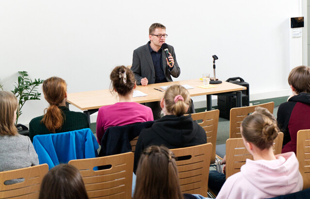 A man sits in a room at a table and speaks to people sitting on chairs in front of him. The audience can only be seen from behind.