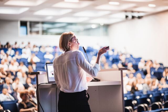 Woman speaker giving a talk at a business conference. Blurry people in the conference hall audience. Business and entrepreneurship event.