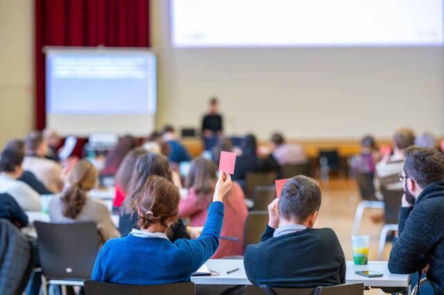 Blurred image of a speaker and audience. Two people are holding up red cards.