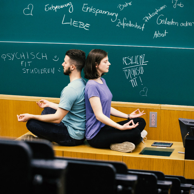 Two students sit cross-legged back to back on the desk in front of the blackboard in the lecture hall, in a meditating arm position, and have their eyes closed.