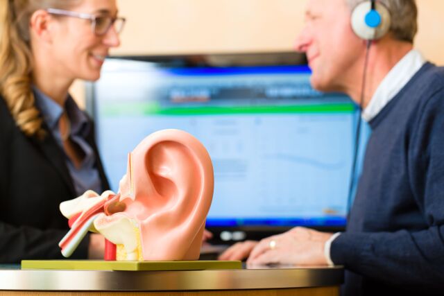 The photo shows a man and a woman sitting opposite each other. The man is wearing headphones and taking a hearing test. In the foreground is a model of an ear. 