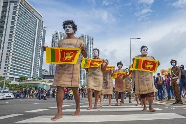 6 people with white painted faces. Each is holding a small Sri Lankan flag. They are walking on a street during a demonstration. Next to them a policeman. Skyscrapers in the background.
