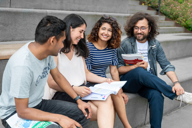 A group of four people are sitting over books and talking. In the corner of the picture, the words “Apply for funding now!” are written in white on a blue background.