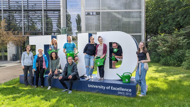 Giant letters spelling out TUD on a lawn. The photo shows a group of people holding watering cans, distributed between the three letters.
