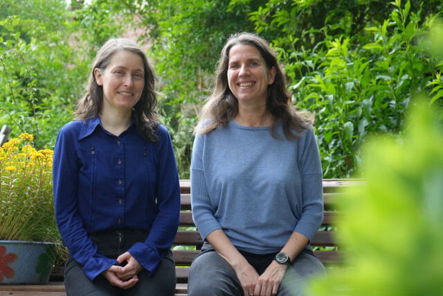 Two women sit on a bench in a garden setting, smiling at the camera. Green foliage in the background, yellow flowers in a pot on the left.