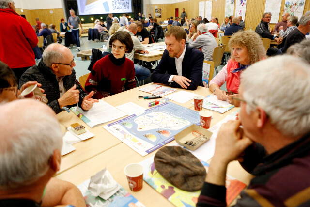 The photo shows people of different ages at a table in conversation. There are pens and paper materials on the table.