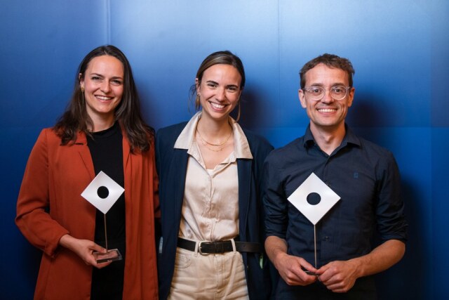 Photo: 3 people in front of a blue background. On the left Dr. Hannah Muti and on the right Prof. Simon Meier-Vieracker, each holding a prize. In the middle Dr. Chiara Löffler.