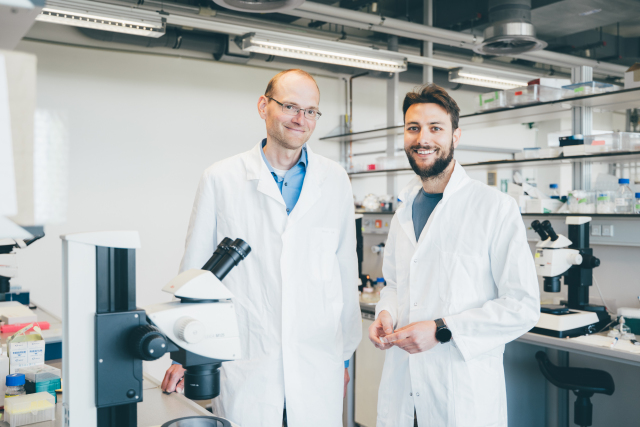 A photo. A bright laboratory with white walls and ceiling lights. In the center, two men wearing white lab coats are standing side by side and smiling at the camera. Behind them, there are laboratory benches with microscopes and various lab supplies.