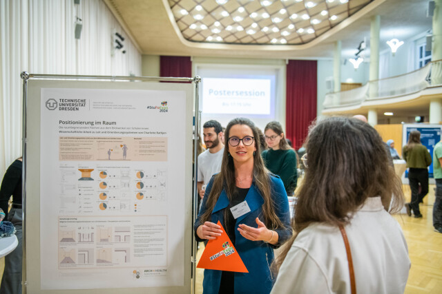 A participant at StuFoExpo 2024 explains her poster to viewers during the poster walk. The poster deals with “positioning in space.” The participant is holding an orange paper triangle in her hand.