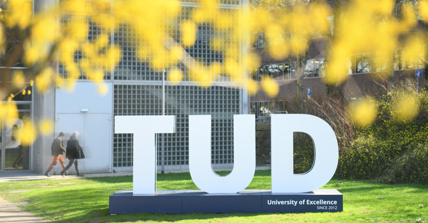 Large white 3D 'TUD' letters on a dark base with the text 'University of Excellence SINCE 2012', on a campus lawn. University buildings and yellow spring blossoms are in the background.